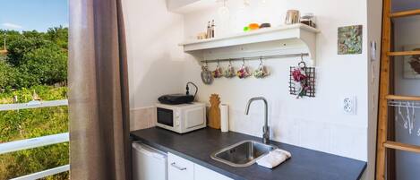 A compact kitchen with a microwave, sink, and white cabinets. A shelf above the sink holds decorative items and a roll of paper towels.