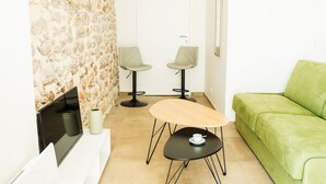 Een moderne woonkamer met een stenen muur, twee grijze stoelen, een groene bank, een klein rond tafeltje en een flatscreen televisie.