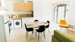 Een compacte keuken met een eethoek, een wasmachine, en een lichte, ruime kamer met een groot raam en een gele stoel.