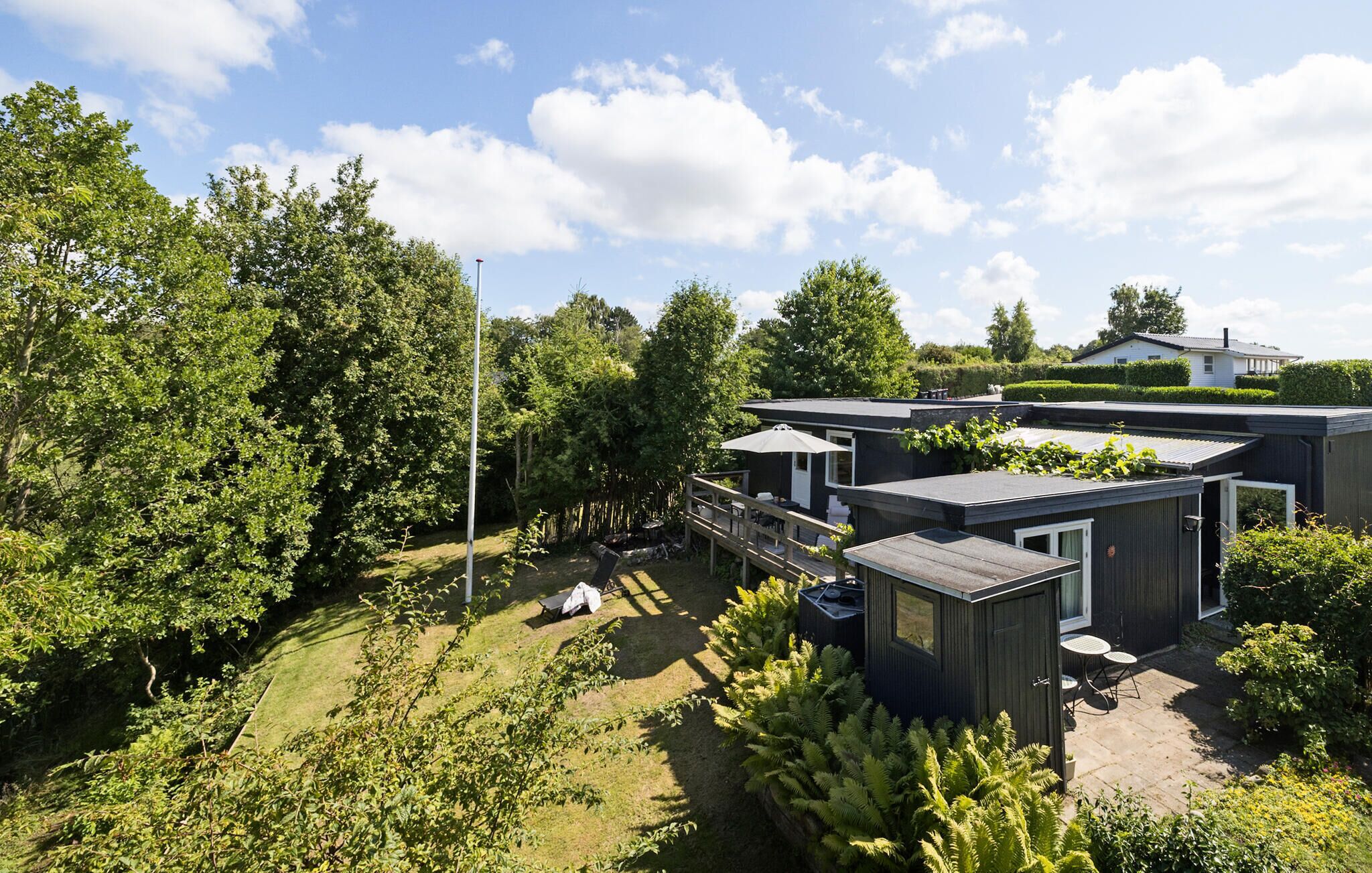Ein moderner Außenbereich mit einer Holzterrasse, einer kleinen Sitzecke und einem Weg zu einem Gebäude mit dunkler Fassade und großen Fenstern.