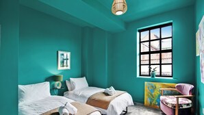 Une chambre d’hôtel aux couleurs turquoise, avec deux lits, une fenêtre donnant sur un paysage agréable, un fauteuil et un tableau au mur.