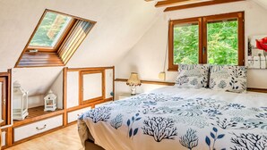 A bedroom with a sloped ceiling, a skylight, a bed with a floral bedspread, two windows with a view of greenery, and wooden furniture.