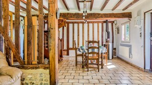 A rustic interior with exposed wooden beams, a tiled floor, a dining table with chairs, and a staircase leading to an upper level.
