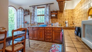 Eine Küche mit Holzschränken, einer Fliesenküchenrückwand, einem Fenster mit Vorhängen und einem Essbereich mit Stuhl und Tisch.