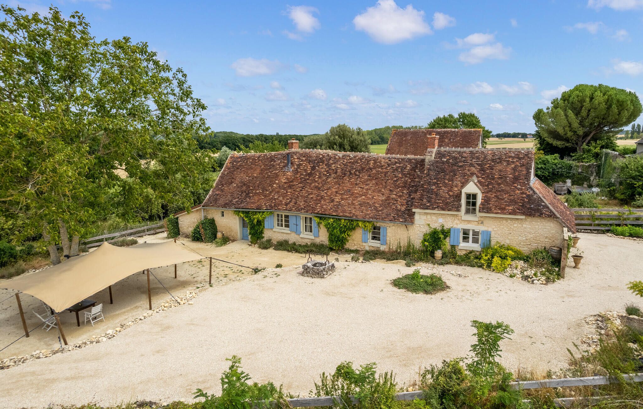 A traditional stone house with a tiled roof, surrounded by a gravel yard and trees.