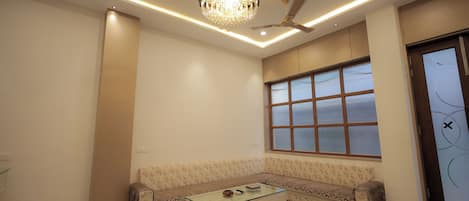 Una sala de estar moderna con un sofá seccional, una mesa de centro de vidrio, una araña, iluminación empotrada en el techo, un ventilador de techo y un ventanal con persianas.