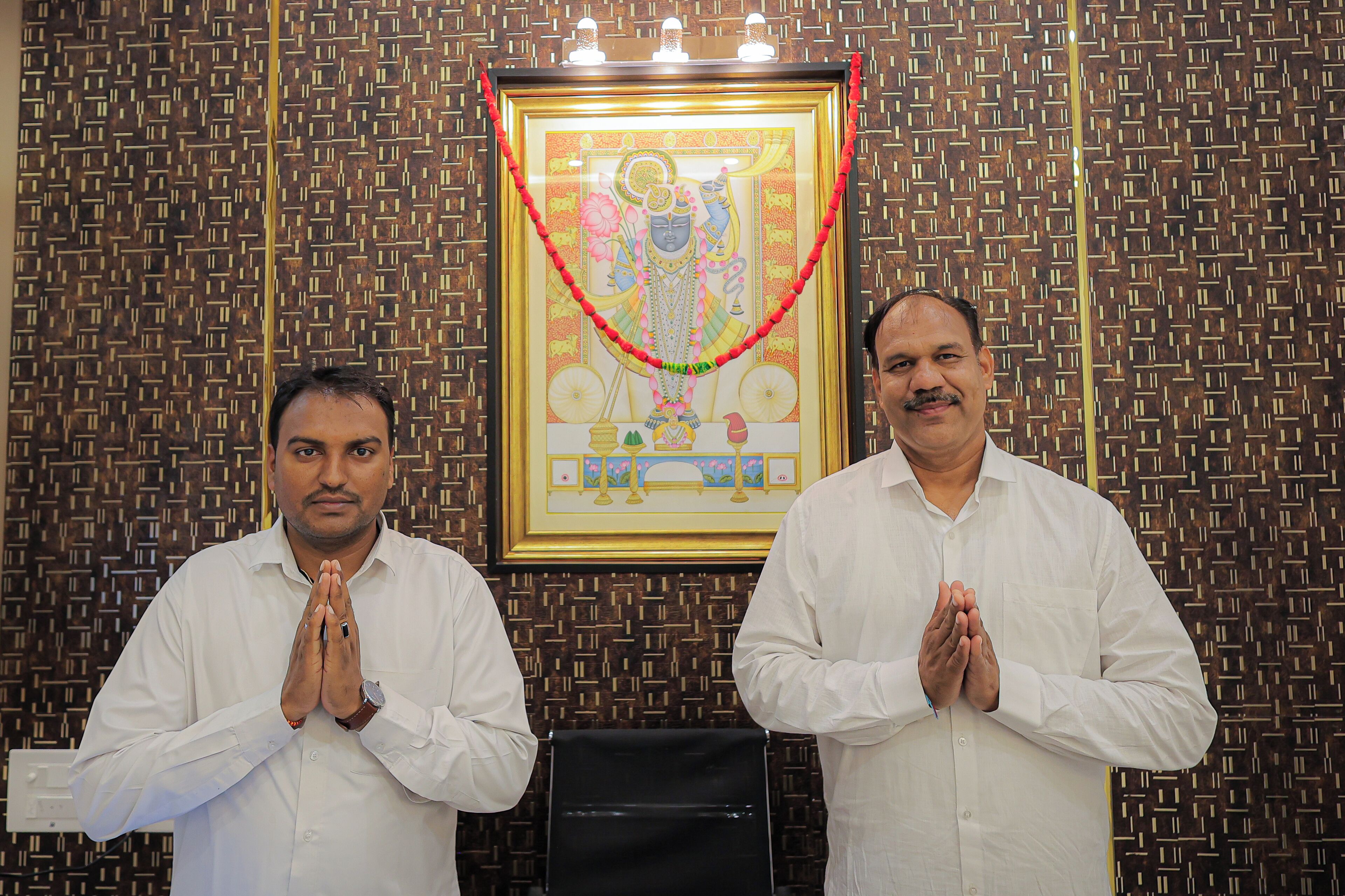 Two men in white shirts standing in front of a patterned wall with a framed religious painting above them.