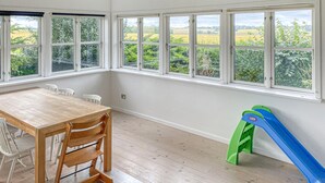 Een ruime kamer met een houten tafel en stoelen, grote ramen met uitzicht op groen, en een glijbaan voor kinderen.