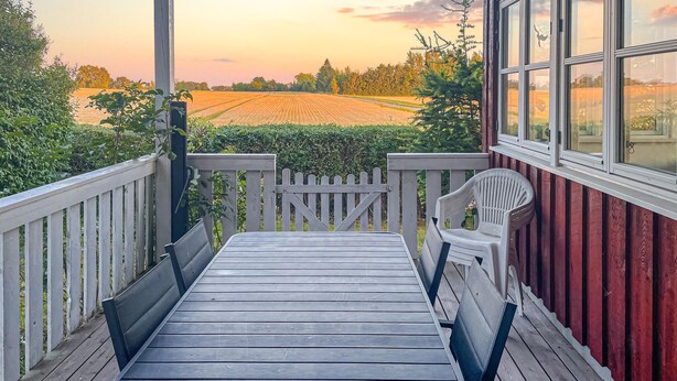 Een houten terras met een witte reling, een witte stoel en uitzicht op een veld en bomen bij zonsondergang.