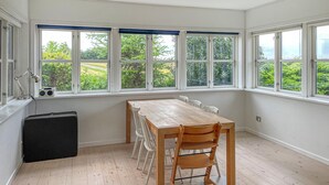 Een kamer met een houten tafel en stoelen, grote ramen met uitzicht op groen, en een zwarte speaker op de vloer.