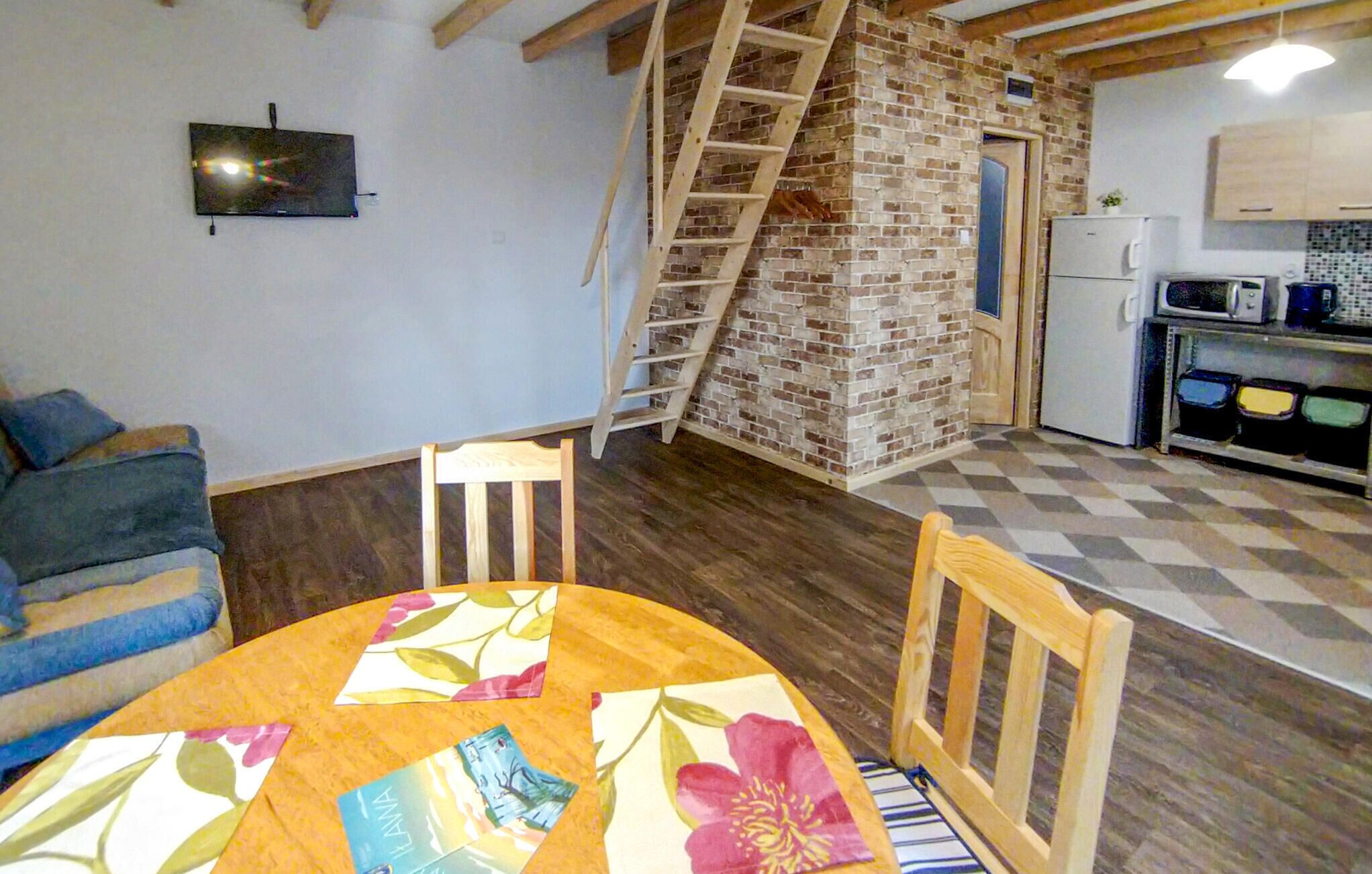 A room with a wooden ceiling, a brick wall, a ladder, a sofa, a table with a floral tablecloth, and a kitchen area with cabinets and a refrigerator.