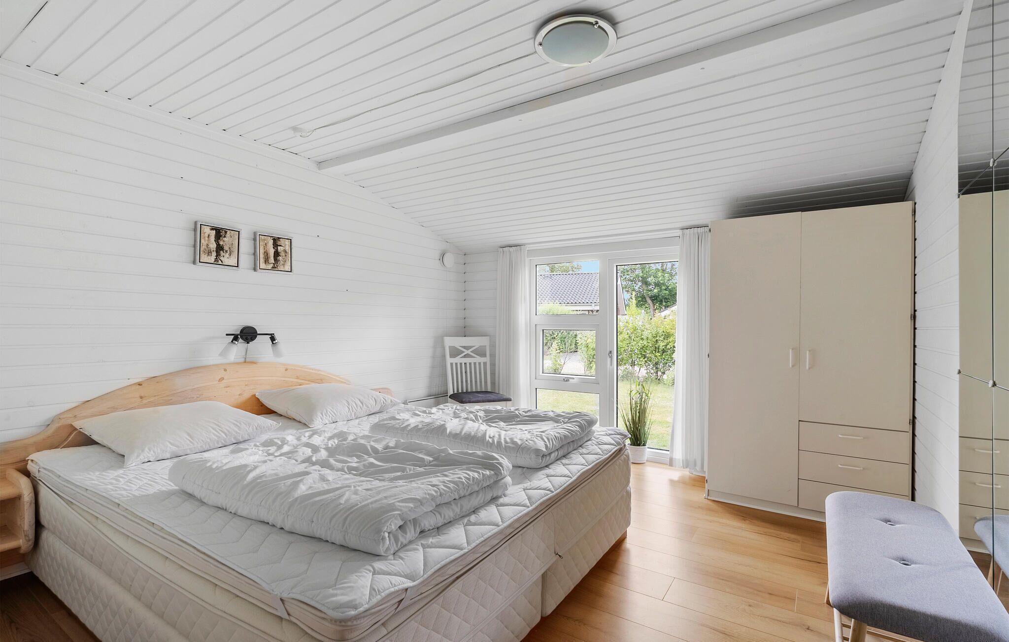 A bedroom with a wooden ceiling, a bed with white bedding, a bedside table, a chair, and a wardrobe.