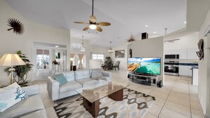 Ein geräumiger Wohnbereich mit einem weißen Sofa, einem Glastisch, einem Deckenventilator, einem Flachbildfernseher und einer Küche mit weißen Schränken und Geräten.