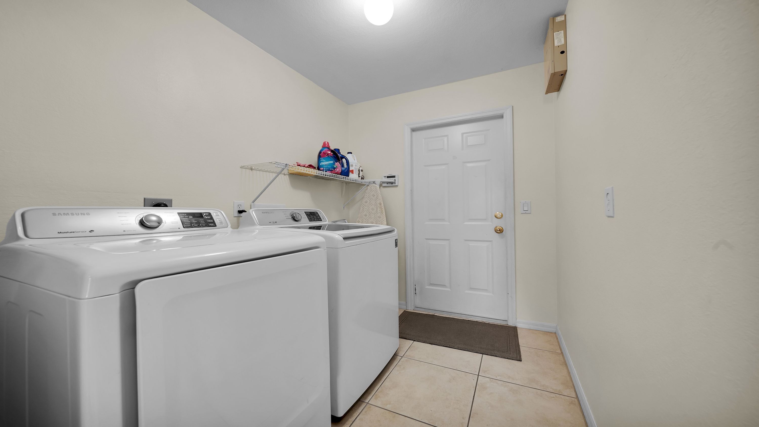 Una sala de bugaderia amb dues rentadores i assecadores frontals blanques, una estant amb productes de neteja i una porta amb felpuda.
