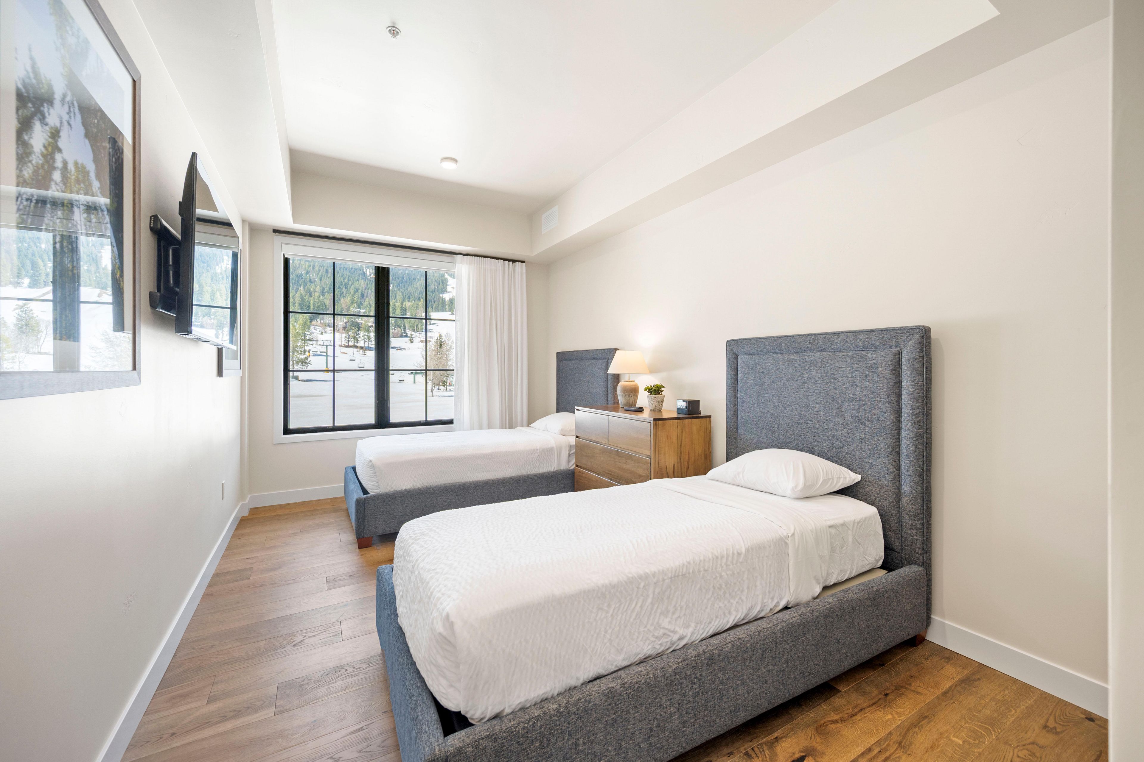 A hotel room with two beds, a wooden floor, a large window with a view, and a bedside table with a lamp.