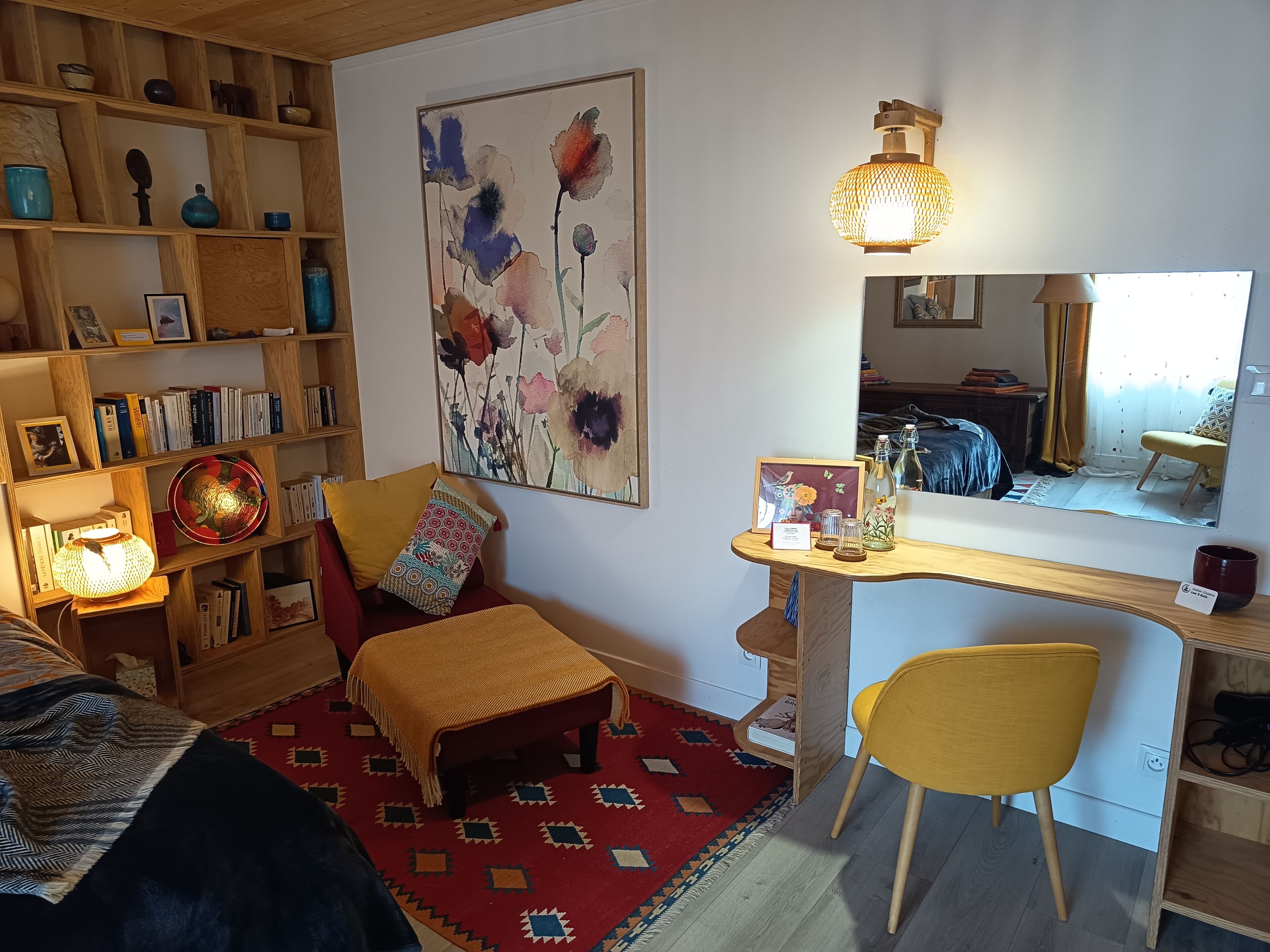 A cozy room with a wooden bookshelf, a desk with a chair, a red rug, and a large floral painting on the wall.