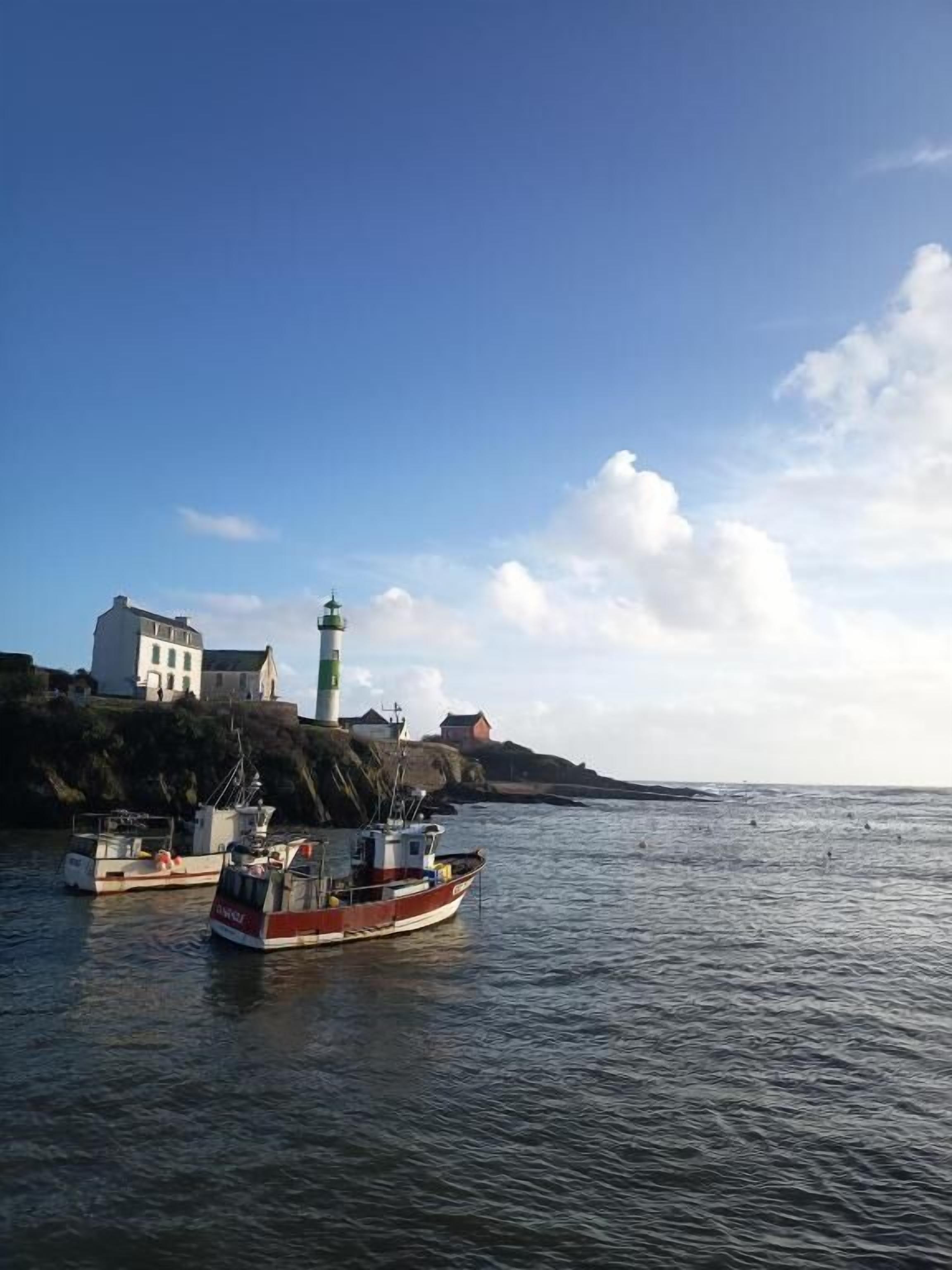 A coastal scene with a lighthouse, a white building, and two boats on calm waters.