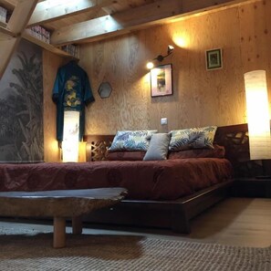 A cozy bedroom with a wooden ceiling, a bed with patterned bedding, a hanging garment, and wall-mounted lighting. - Les 3 Koïs (Riec-sur-Bélon)