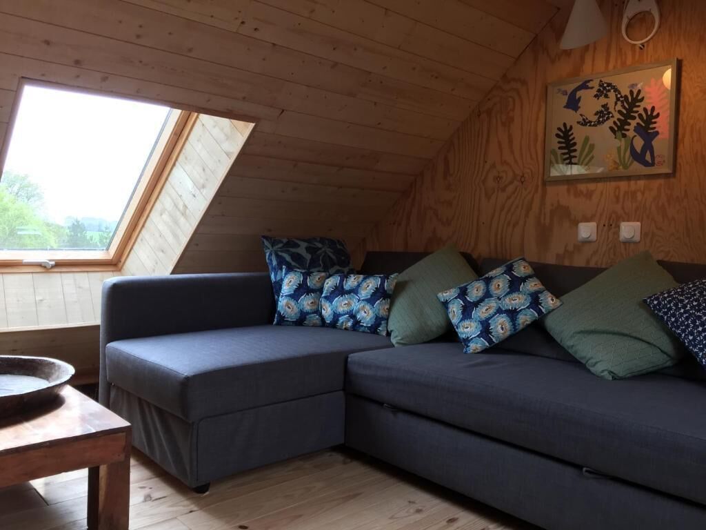 A cozy room with a wooden ceiling, a large window, a dark blue sectional sofa with patterned cushions, and a small wooden table.