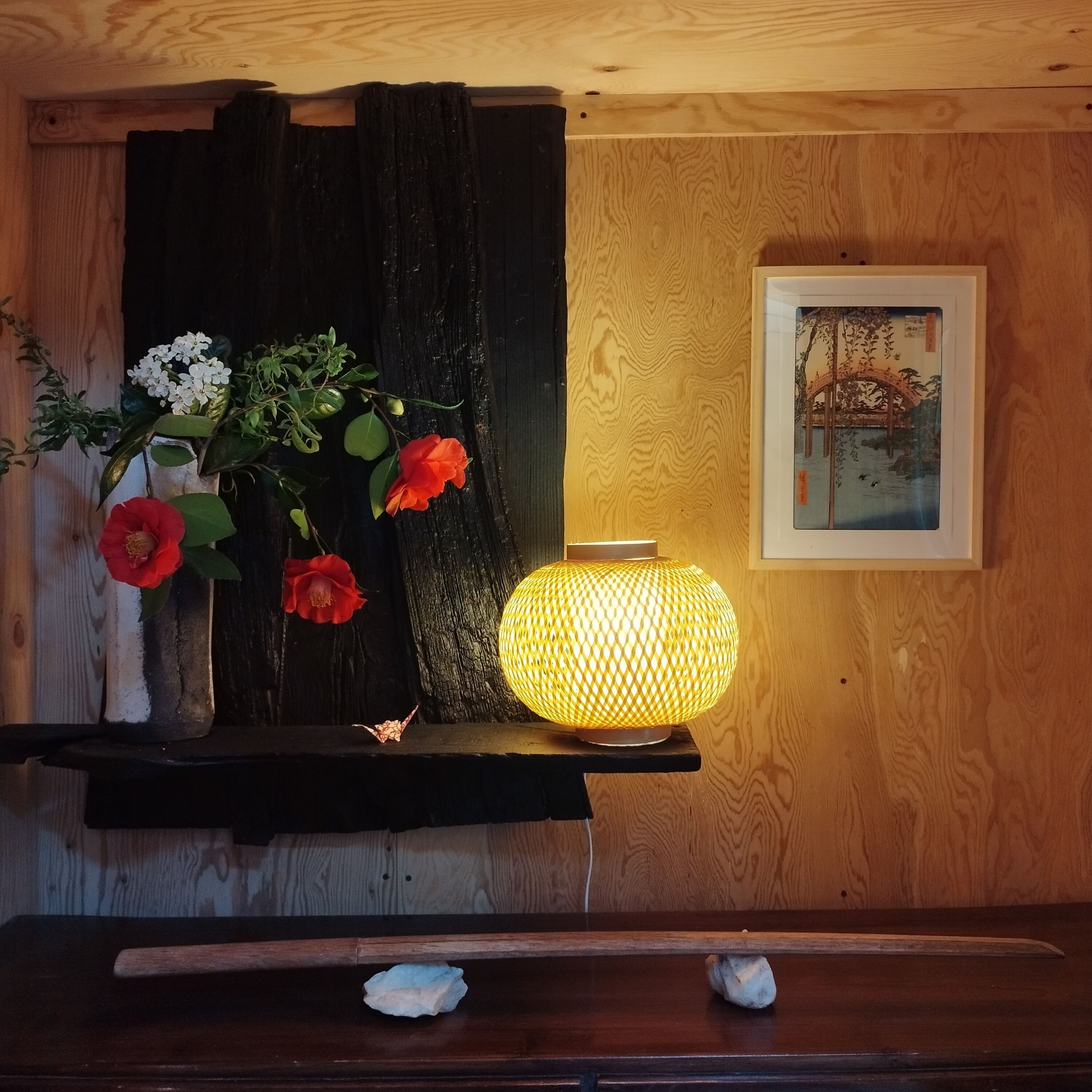 En hylla i trä med en vas med blommor, en tänd lampa och en inramad tavla på väggen.