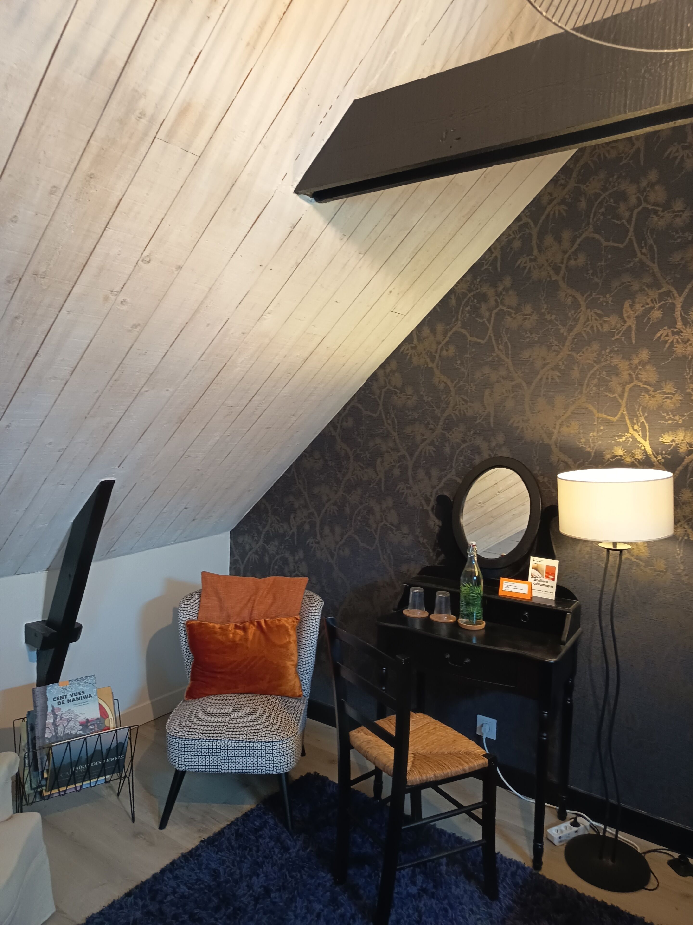A room with a sloped ceiling, a black desk with a mirror and lamp, a patterned chair, and a blue rug.