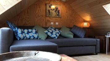 A cozy loft-style room with a wooden ceiling, a gray sofa with patterned cushions, a wooden coffee table, and a wall-mounted light fixture.