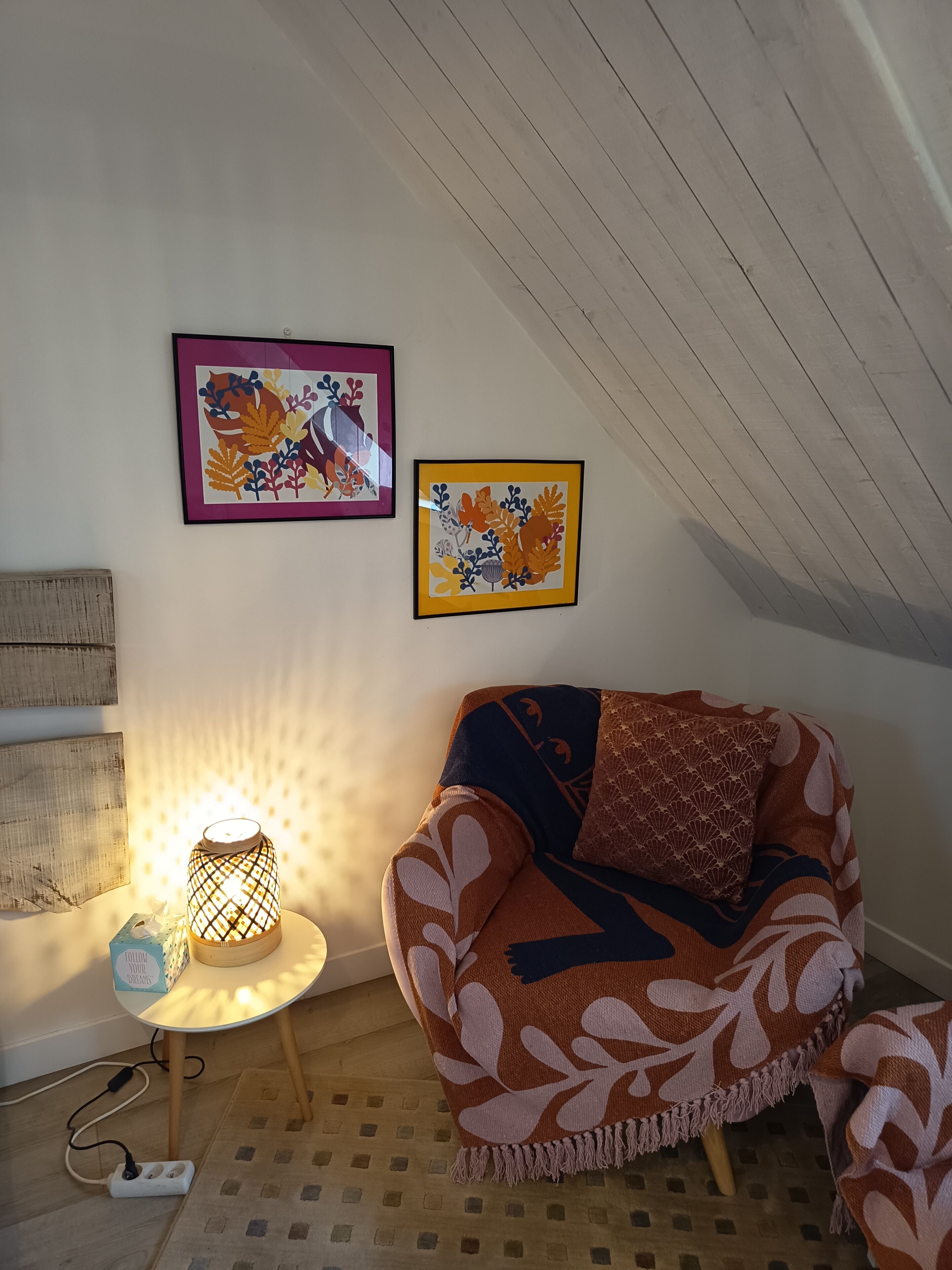 A cozy room with a sloped ceiling, a cushioned armchair, a small table with a lamp, and two framed artworks on the wall.