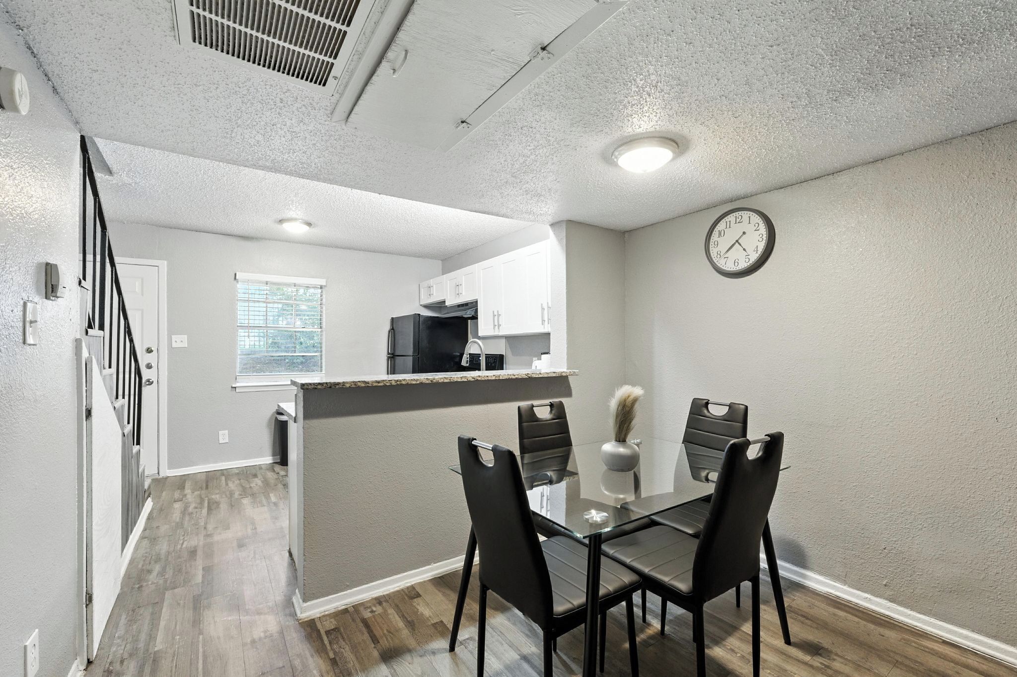 Una cocina moderna con una isla central, un comedor con mesa de vidrio y sillas negras, y un reloj en la pared.
