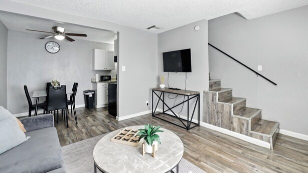 A modern living room with a grey sofa, a round coffee table, a dining area with a table and chairs, a kitchen area, and a staircase with a TV mounted on the wall.