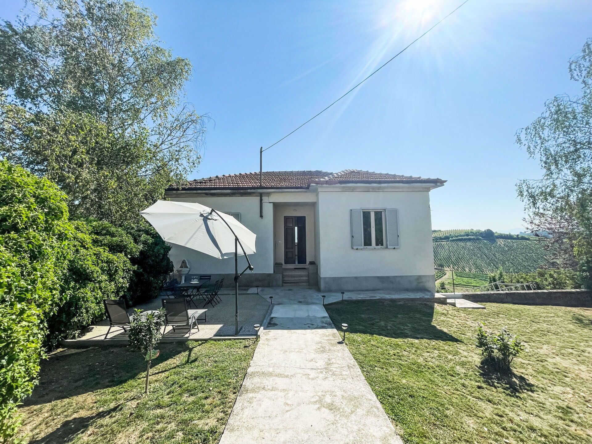 A small house with a tiled roof, a door, and a window. There is a patio with a table and chairs, and a large umbrella.