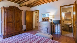 Un dormitorio con un armario de madera grande, una cama con colcha estampada, un escritorio con lámpara y un baño visible a través de una puerta abierta.