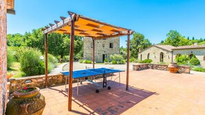 Una mesa de ping pong al aire libre bajo una pérgola de madera, rodeada de edificios de piedra y vegetación.