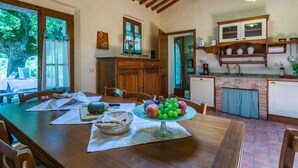 Un área de comedor con una mesa de madera, sillas y un centro de mesa con frutas. Se observa también una cocina con armarios y un lavamanos.