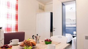 A hotel room with a dining table set with food and drinks, a bed with a blue headboard, and a kitchenette area. - Beautiful apartment in villa for 5 guests with pool, A/C, WIFI, terrace and pets allowed (Capalbio)