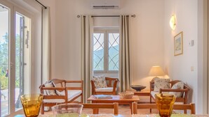 A dining area with a table set for two, featuring a plate of fruit, a bowl of tomatoes, and two glasses. There are two wooden chairs and a sofa with cushions. A window with curtains is visible in the background. - Stunning private villa for 4 people with WIFI, A/C, TV, patio, pets allowed and panoramic view (Olbia)