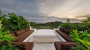 Une terrasse sur le toit, dotée d’un espace salon avec des canapés marron et un pouf blanc, entourée d’une végétation luxuriante et offrant une vue sur une étendue d’eau.