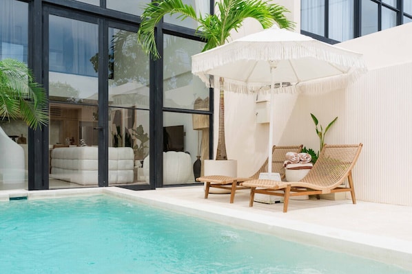 Zona de piscina con una sombrilla blanca, dos sillones de mimbre y vistas a un spa a través de amplios ventanales.