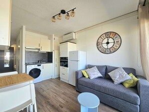 Um espaço de estar compacto com cozinha americana, um sofá cinza com almofadas estampadas, um relógio de parede redondo e uma pequena mesa redonda.
