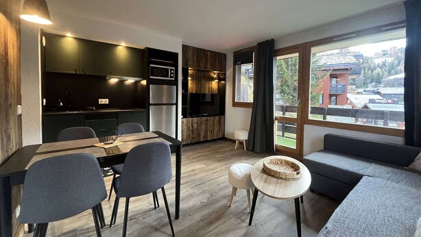 Una cocina moderna con armarios oscuros, microondas y refrigerador. Hay una zona de comedor con una mesa y sillas, y un ventanal con vistas a una estación de esquí.