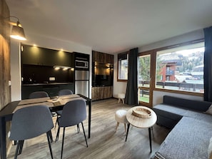 Una cocina moderna con armarios oscuros, microondas y refrigerador. Hay una zona de comedor con una mesa y sillas, y un ventanal con vistas a una estación de esquí.