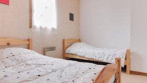 Un dormitorio con dos camas de madera, una más grande y otra más pequeña, ambas con ropa de cama blanca con estampados de estrellas. Hay una ventana que da a vistas exteriores y un pequeño calentador en el suelo.