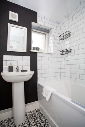 A bathroom with a white pedestal sink, a mirror, a window with frosted glass, a white bathtub, and a black and white patterned floor. - Vacant Nests | The Darwin | Stylish | Wifi (Hartlepool)