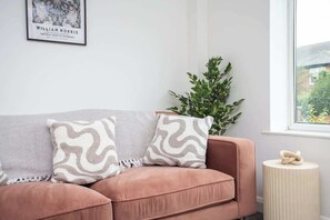 A room with a pink sofa, patterned cushions, a framed picture on the wall, a potted plant, and a small round table. - Vacant Nests | The Darwin | Stylish | Wifi (Hartlepool)