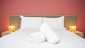 En pænt redt seng med hvide sengetøj, to puder og et sammenrullet håndklæde. Der er to sengeborde med lamper på hver side af sengen.