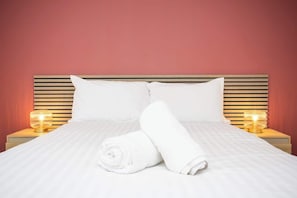 A neatly made bed with white linens, two pillows, and a rolled towel. There are two bedside lamps on either side of the bed.