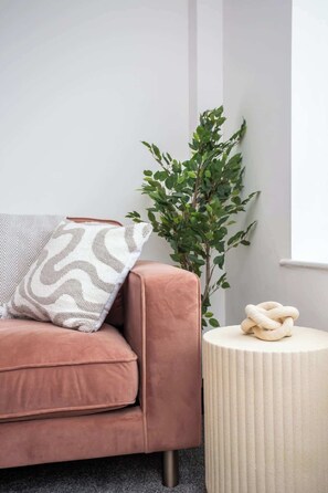 A room with a pink sofa, a patterned pillow, a green plant, and a round side table with a coiled rope.