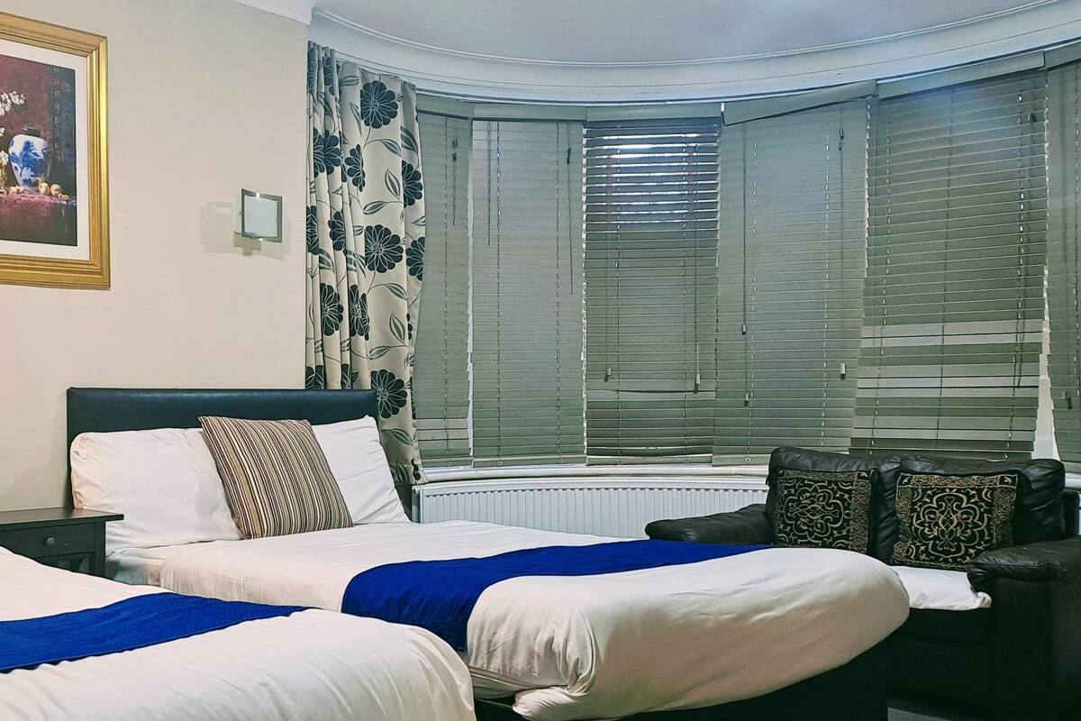 A hotel room with two beds, a nightstand, a framed picture, a window with blinds, and a sofa.