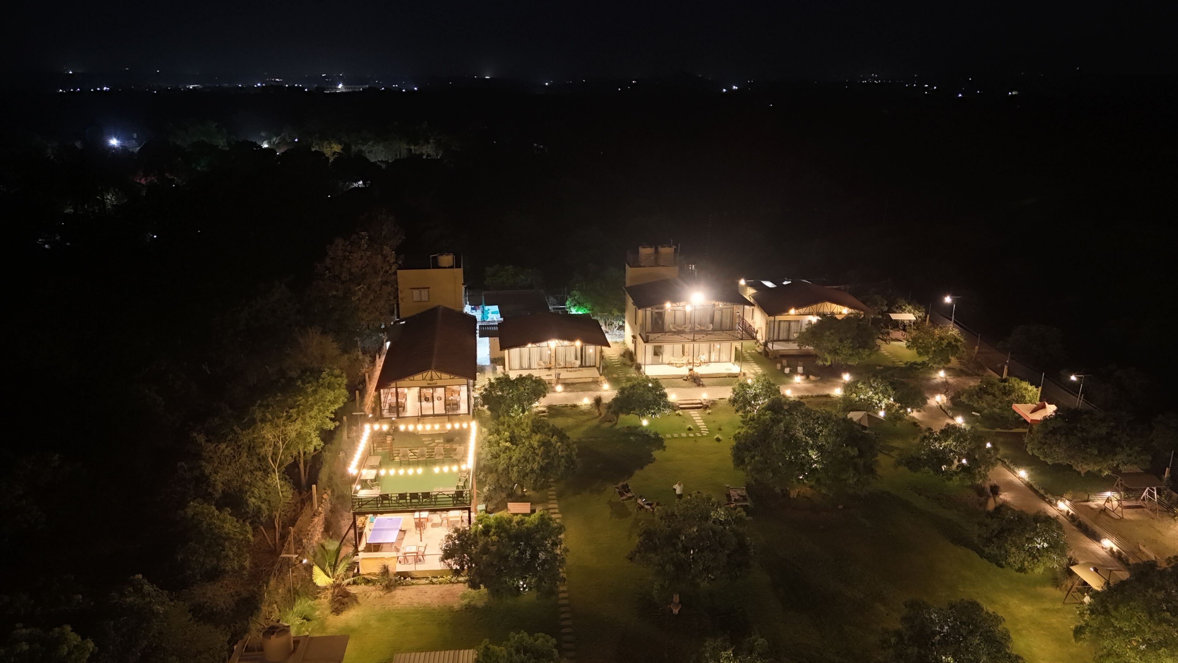 Un resort bien iluminado con varias edificaciones, piscina y jardín ajardinado.