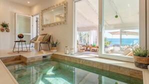 A spacious indoor swimming pool with clear water, a sunlit seating area with a view of the sea, and a modern interior design. - ClickSardinia Villa Tramonto by the beach with path to the sea (sorso)