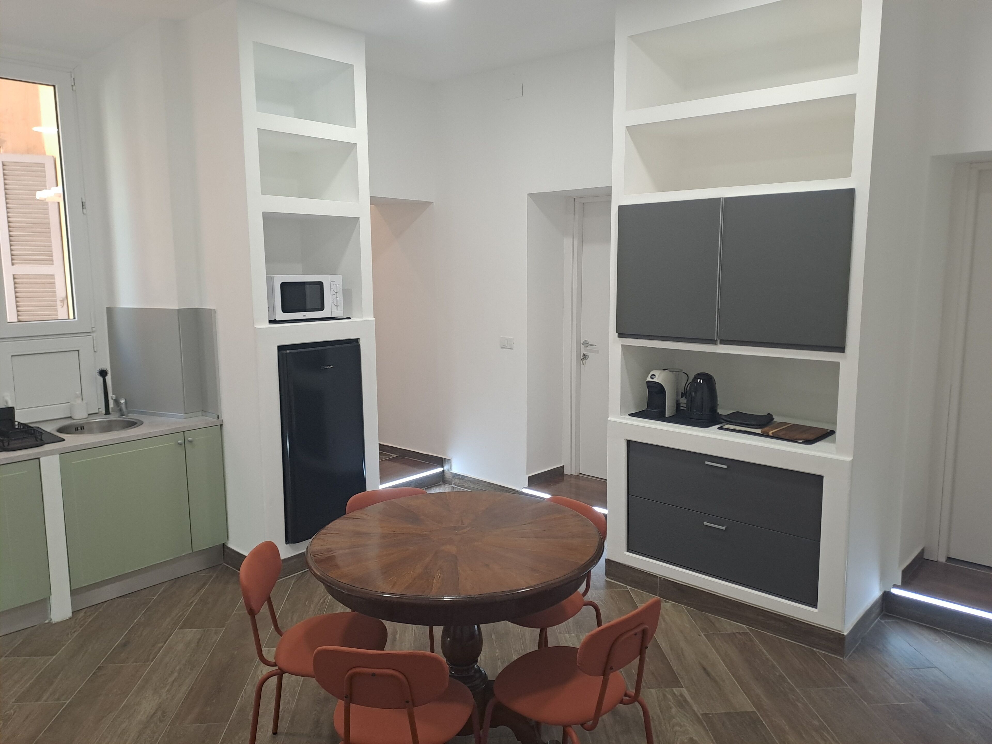 A compact kitchen with a round wooden table and four chairs, a built-in microwave, a small refrigerator, and open shelving with kitchen items.